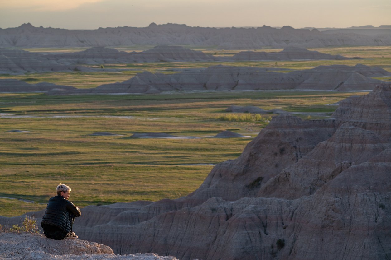 A person wearing a black jacket sits on a rocky cliff, overlooking layered, rugged badlands and green plains stretching into the distance under soft evening light.