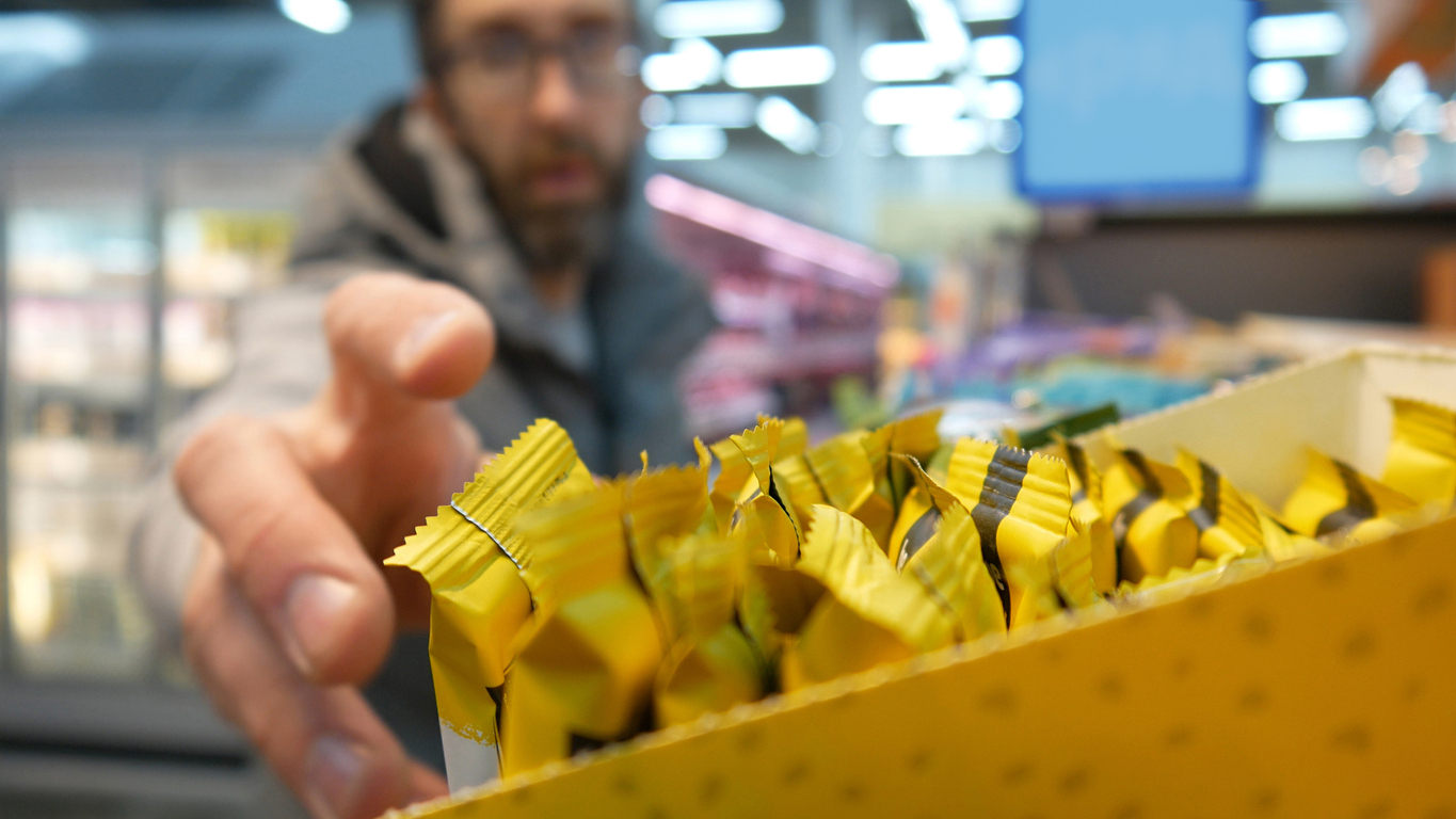 A person reaches for a yellow-wrapped snack bar from a display box in a brightly lit store, with shelves and products blurred in the background.