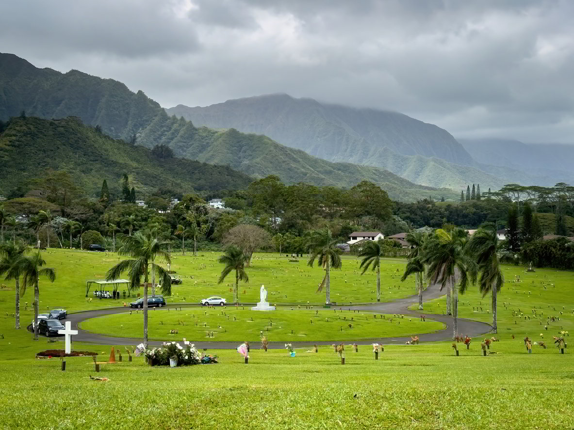 A cemetery with palm trees and a white statue in the center, surrounded by mountains under a cloudy sky. People and cars are scattered across the green grass, with gravestones visible throughout the scene.