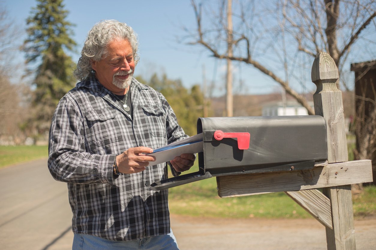 A smiling older man with gray curly hair and a plaid shirt checks his mailbox on a sunny day, holding a few envelopes while standing next to a rural roadside mailbox with the red flag up.