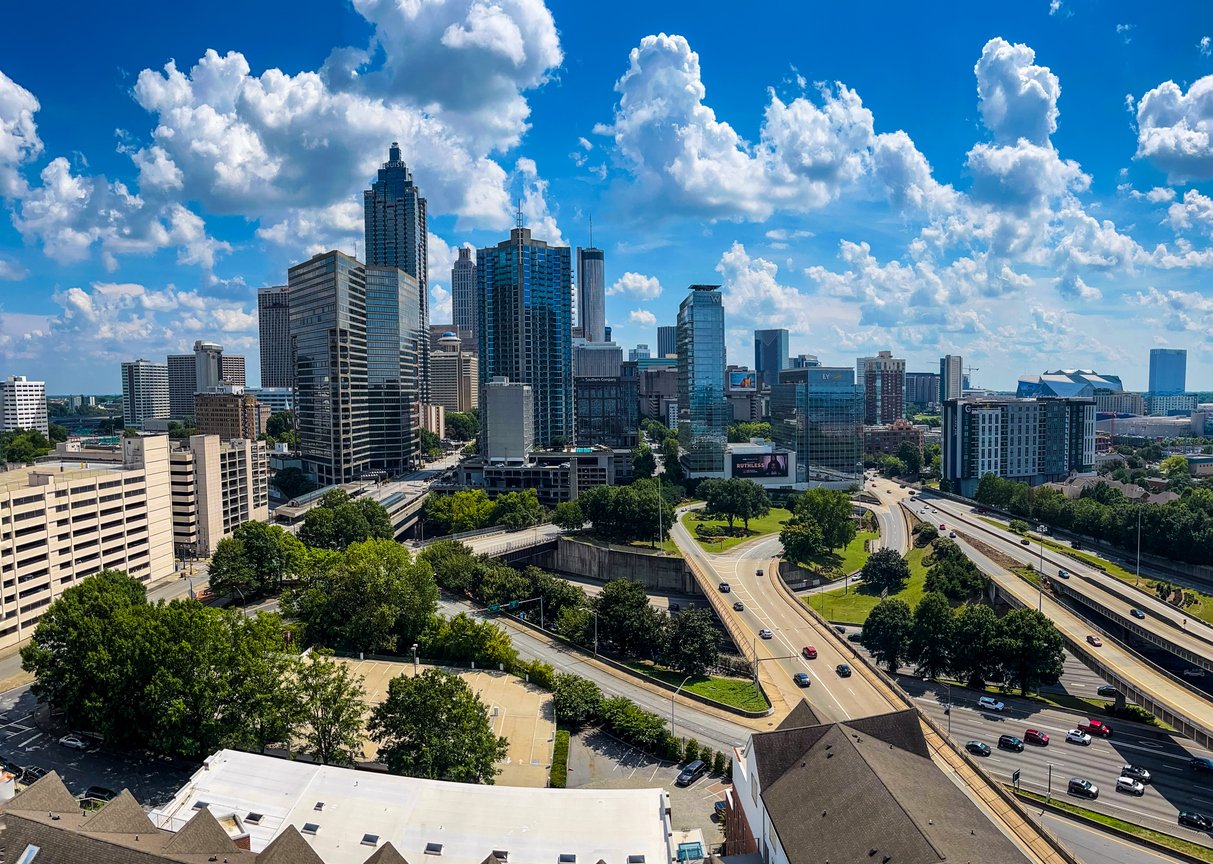A vibrant cityscape of downtown Atlanta, Georgia, with tall skyscrapers, busy highways, green trees, and a bright blue sky dotted with fluffy white clouds.