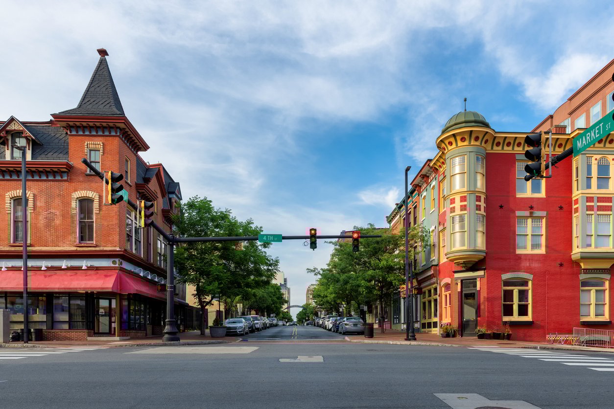 A quiet intersection in a historic downtown area with colorful, ornate buildings on each corner, traffic lights, parked cars, and trees lining the street under a blue sky with wispy clouds.