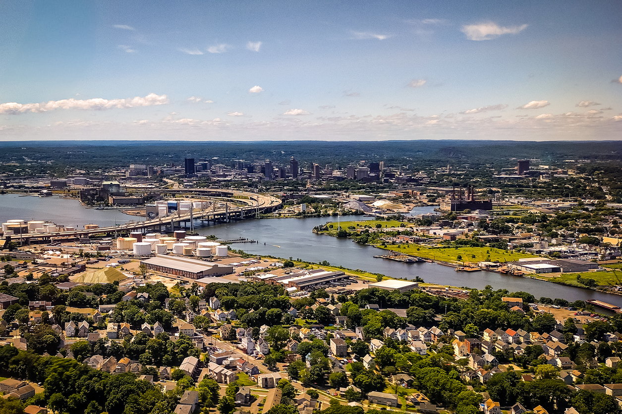 Aerial view of a city with a river running through it, bridges, industrial facilities, residential neighborhoods in the foreground, and a downtown skyline in the background under a mostly clear sky.