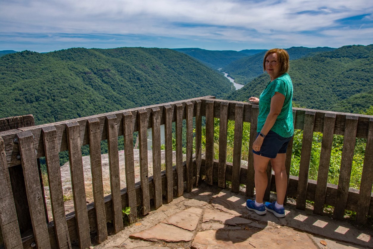 A woman stands on a wooden observation deck overlooking a lush green valley with a winding river below, surrounded by forested hills under a partly cloudy sky.
