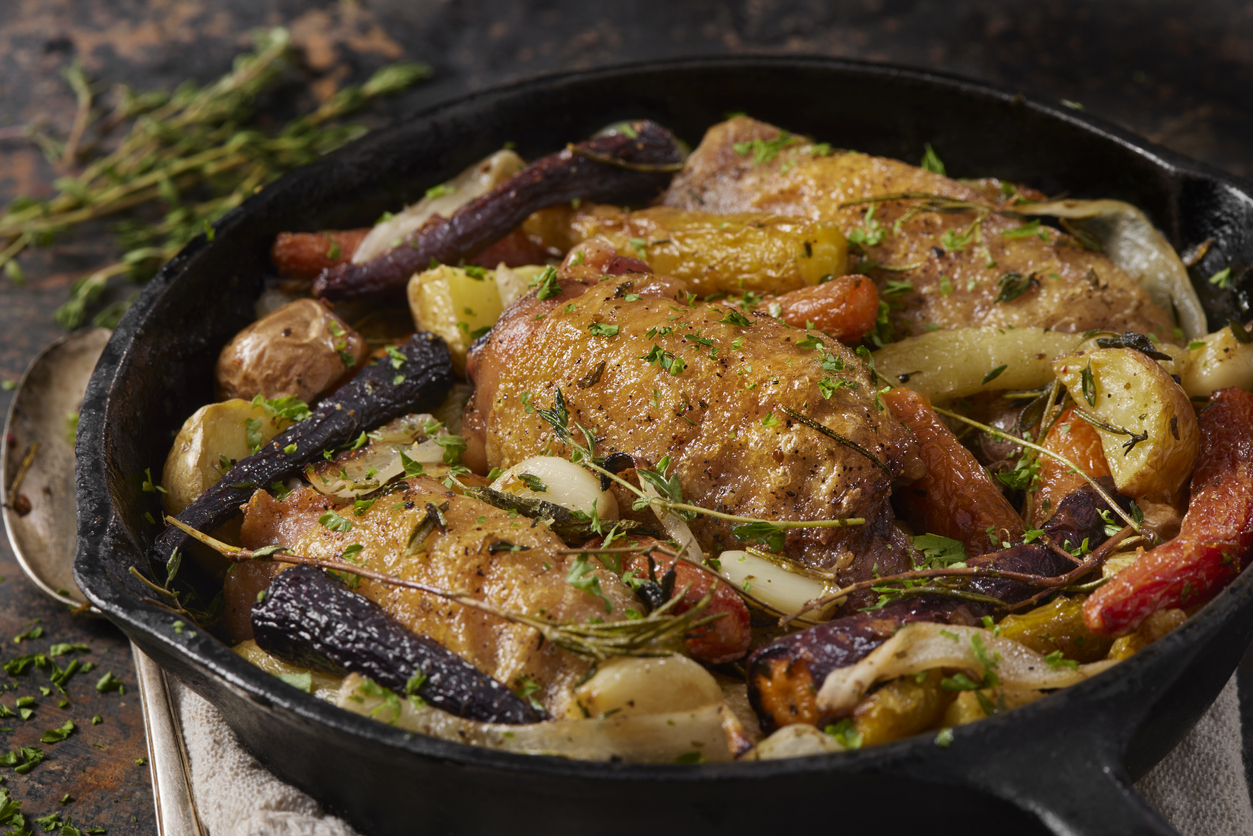 A cast iron skillet filled with roasted chicken thighs, colorful carrots, potatoes, and fresh herbs, garnished with chopped parsley. The dish appears golden brown and savory.