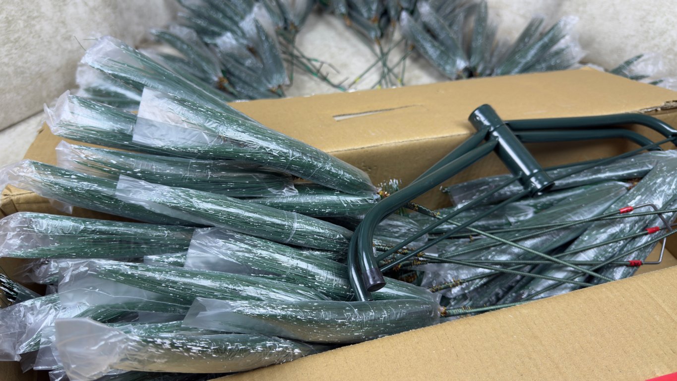 A cardboard box filled with artificial pine branches wrapped in plastic, next to a green metal wreath frame. More pine branches are visible in the background.