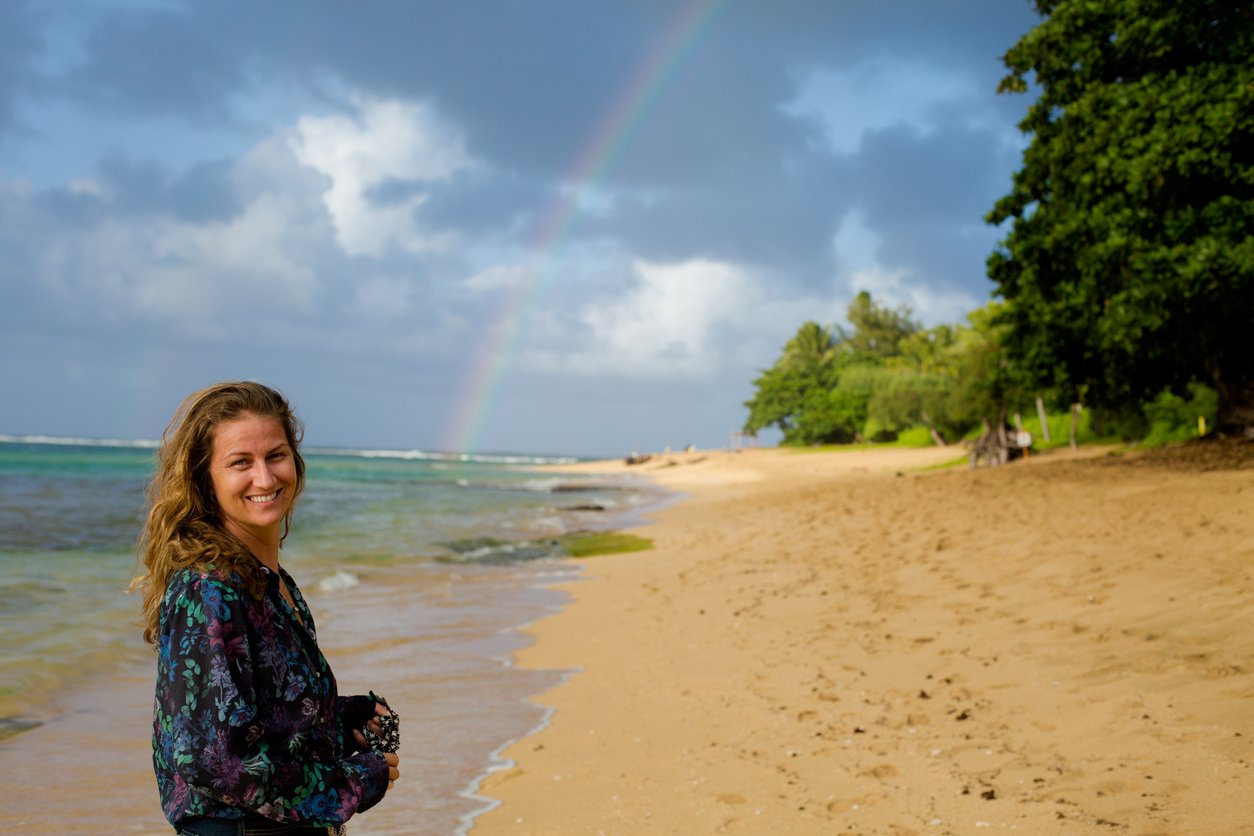 A smiling woman stands on a sandy beach near the water, with lush greenery and trees in the background. A faint rainbow appears in the partly cloudy sky above the shoreline.