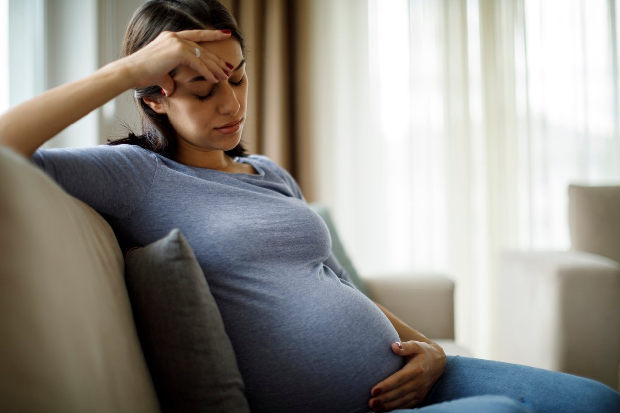 A pregnant woman sits on a couch, holding her belly with one hand and touching her forehead with the other, looking tired or stressed. Light streams through curtains in the background.