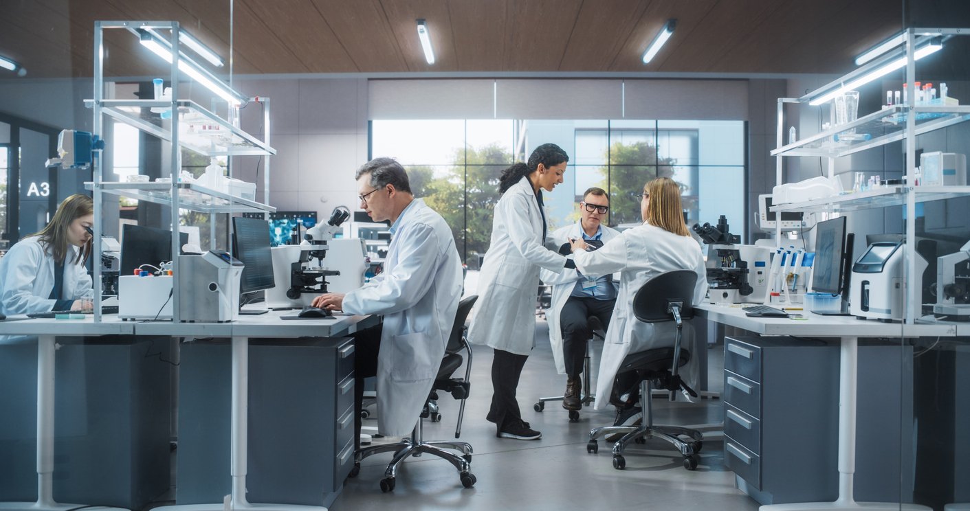 A modern laboratory with five scientists in white lab coats working at desks and lab benches, using computers and scientific equipment, with large windows in the background letting in natural light.