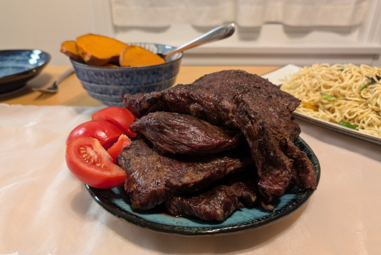 A plate of grilled steak slices stacked with fresh tomato wedges, next to a bowl of roasted pumpkin and a platter of noodles on a dining table.