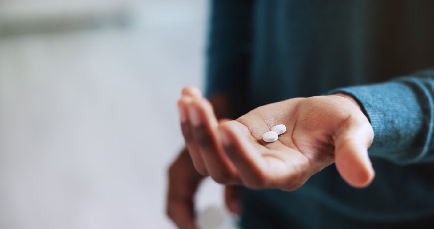 A person wearing a blue long-sleeve shirt holds two white round pills in the palm of their hand, with fingers slightly curled and the background softly blurred.