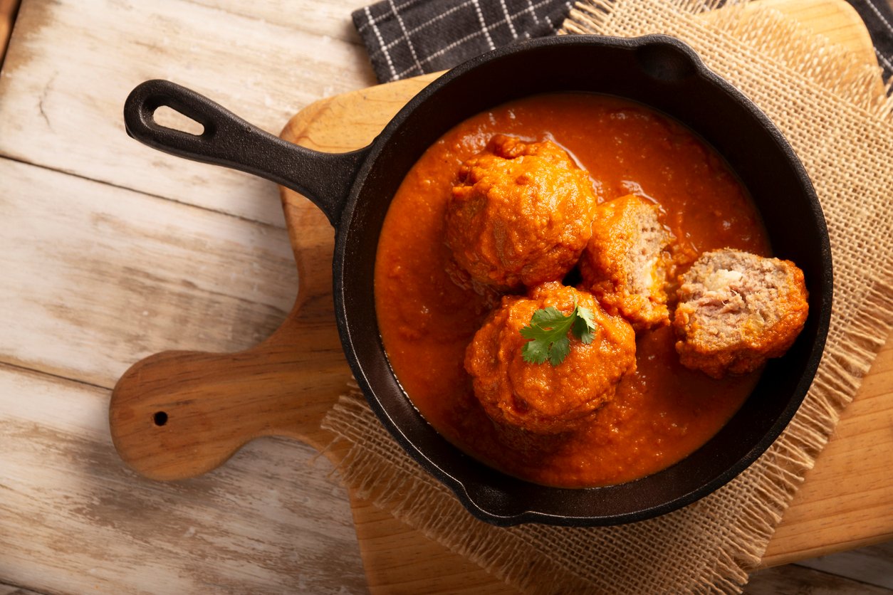 Three meatballs in tomato sauce are served in a black cast iron skillet, garnished with parsley. One meatball is cut open, showing the inside. The skillet sits on a wooden board with burlap and a cloth napkin.