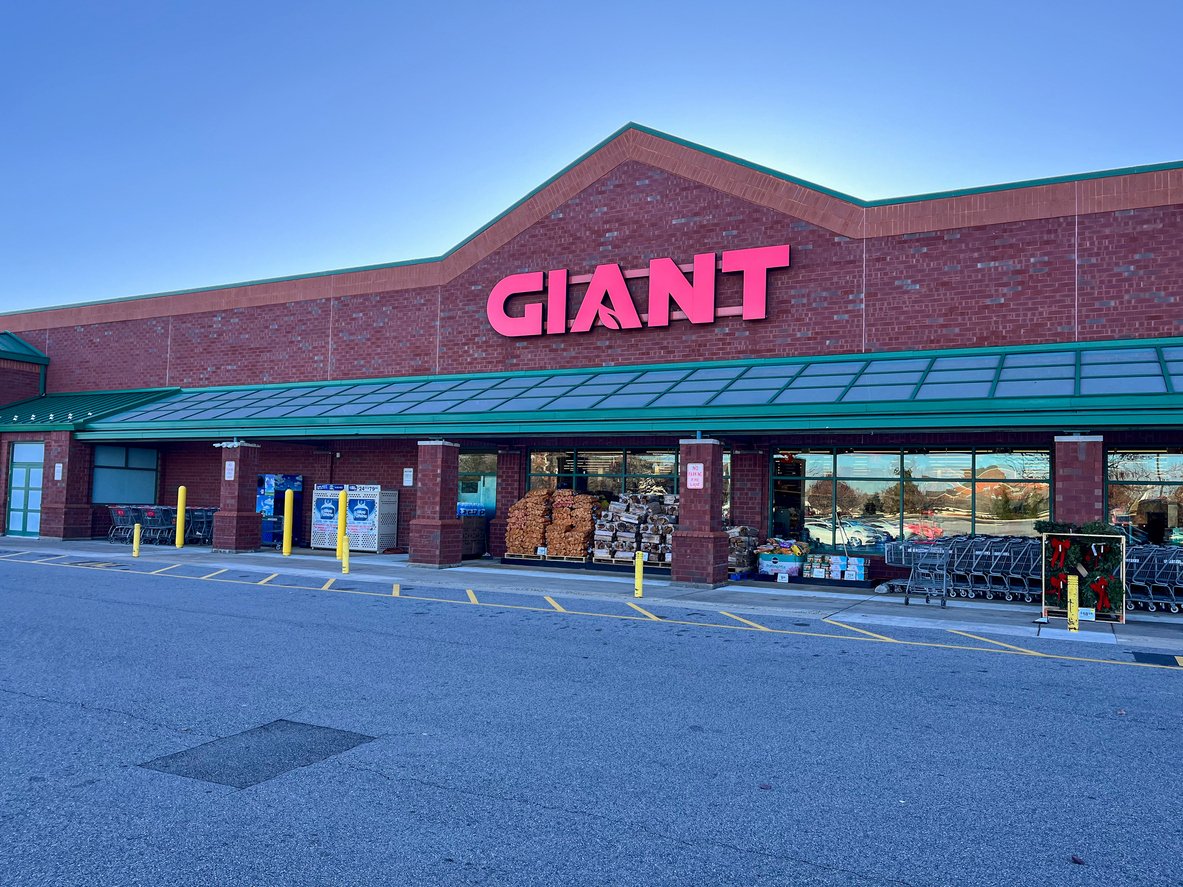 A large brick supermarket with a green awning and a bright red "GIANT" sign above the entrance. Shopping carts and stacked goods are visible outside, with clear blue sky overhead.