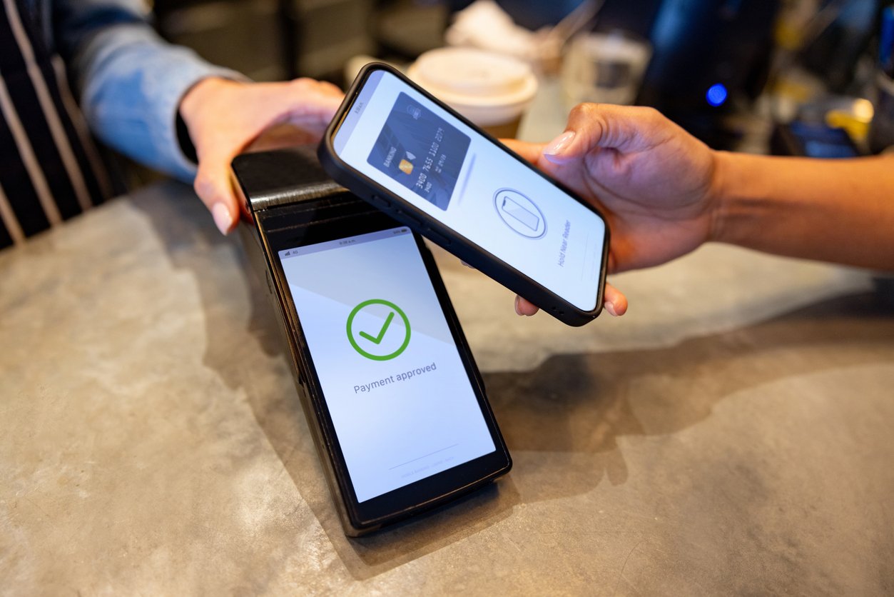 A person holds a smartphone over a card reader, making a contactless payment. The payment terminal screen shows a green checkmark with the message "Payment approved." Another hand holds the terminal.