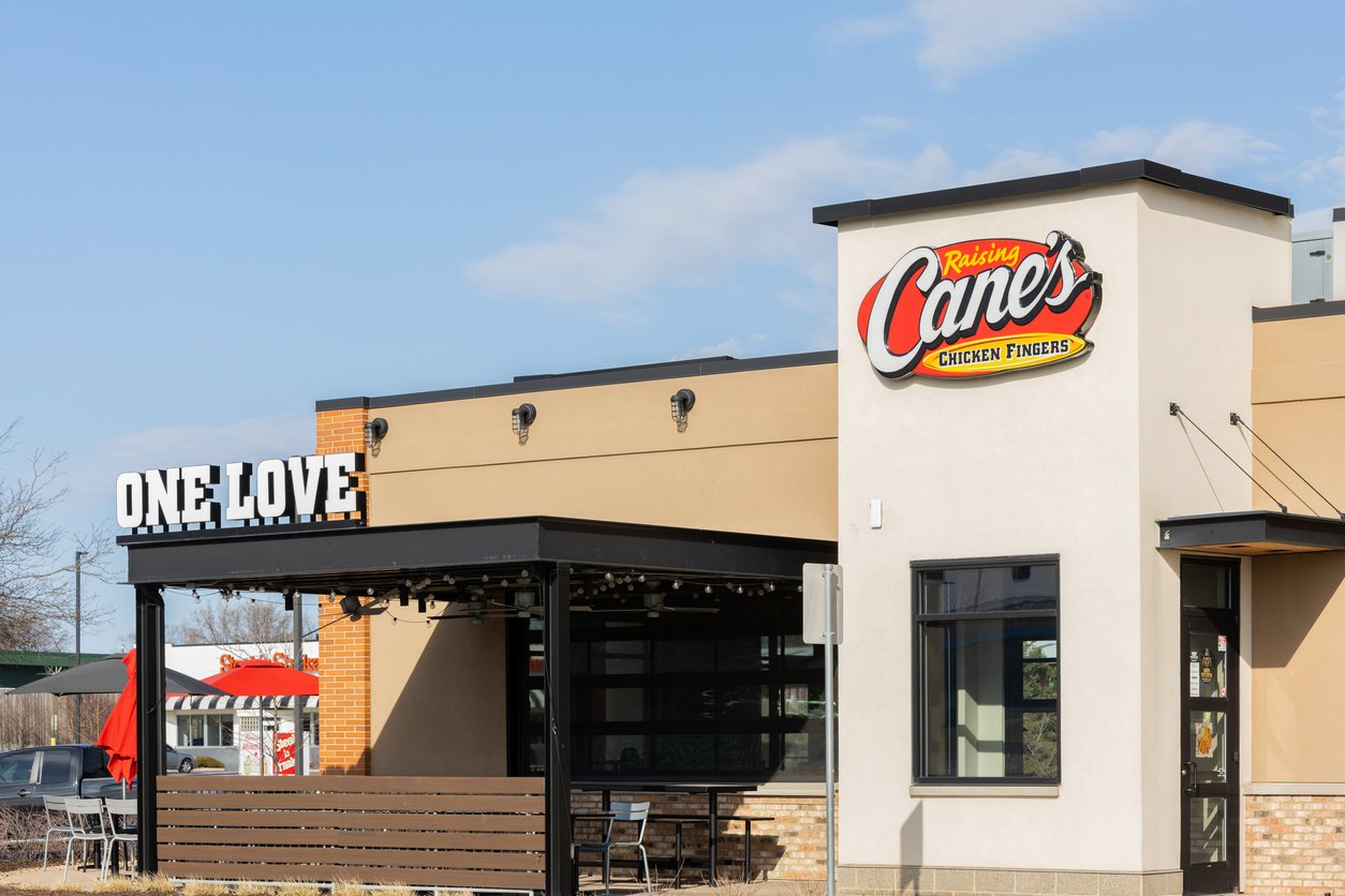 A Raising Cane’s restaurant exterior on a sunny day, featuring the logo and a “ONE LOVE” sign, with outdoor seating and a red umbrella visible in the background.