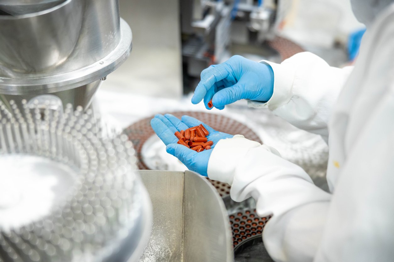 A person wearing blue gloves and a white lab coat holds several red capsules in one hand while working at a pharmaceutical manufacturing facility with machinery and equipment visible.