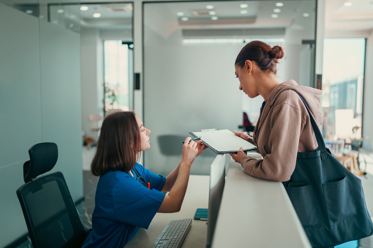 A receptionist in blue scrubs hands a clipboard with forms to a woman standing at a front desk. The woman, carrying a black bag, leans forward to receive the clipboard in a modern, bright office setting.