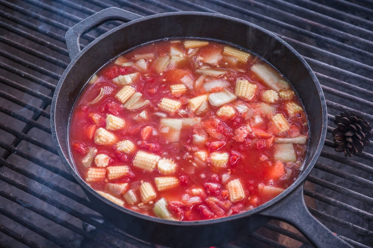 A black cast iron pot sits on a grill, filled with a simmering tomato-based stew containing baby corn, chopped tomatoes, onions, and other vegetables. Steam rises from the pot.
