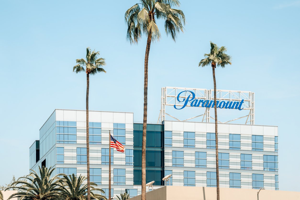 A modern glass building with the "Paramount" sign on top, surrounded by tall palm trees and an American flag in the foreground under a clear sky.