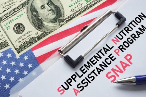 A clipboard with papers labeled “Supplemental Nutrition Assistance Program (SNAP)” sits on an American flag, next to a $100 bill and a blue pen.