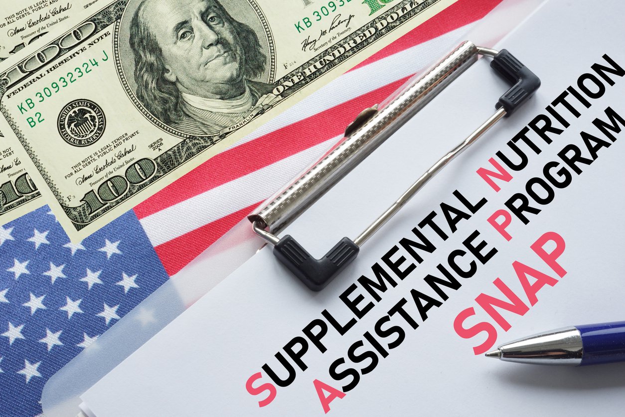 A clipboard with papers labeled “Supplemental Nutrition Assistance Program (SNAP)” sits on an American flag, next to a $100 bill and a blue pen.