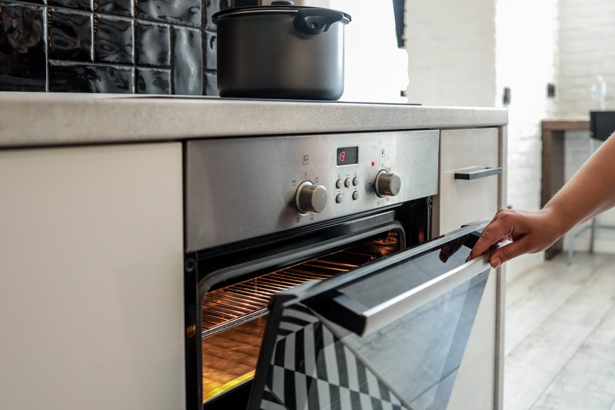 A person’s hand opens an oven door in a modern kitchen, revealing empty oven racks. A black pot sits on the stovetop above. The oven display shows a red digital number and the control knobs are visible.