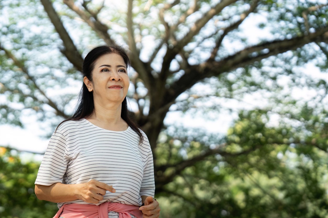 A woman with long dark hair, wearing a striped shirt, stands outdoors smiling with a tree and green foliage in the background. Sunlight filters through the leaves, creating a peaceful, natural setting.