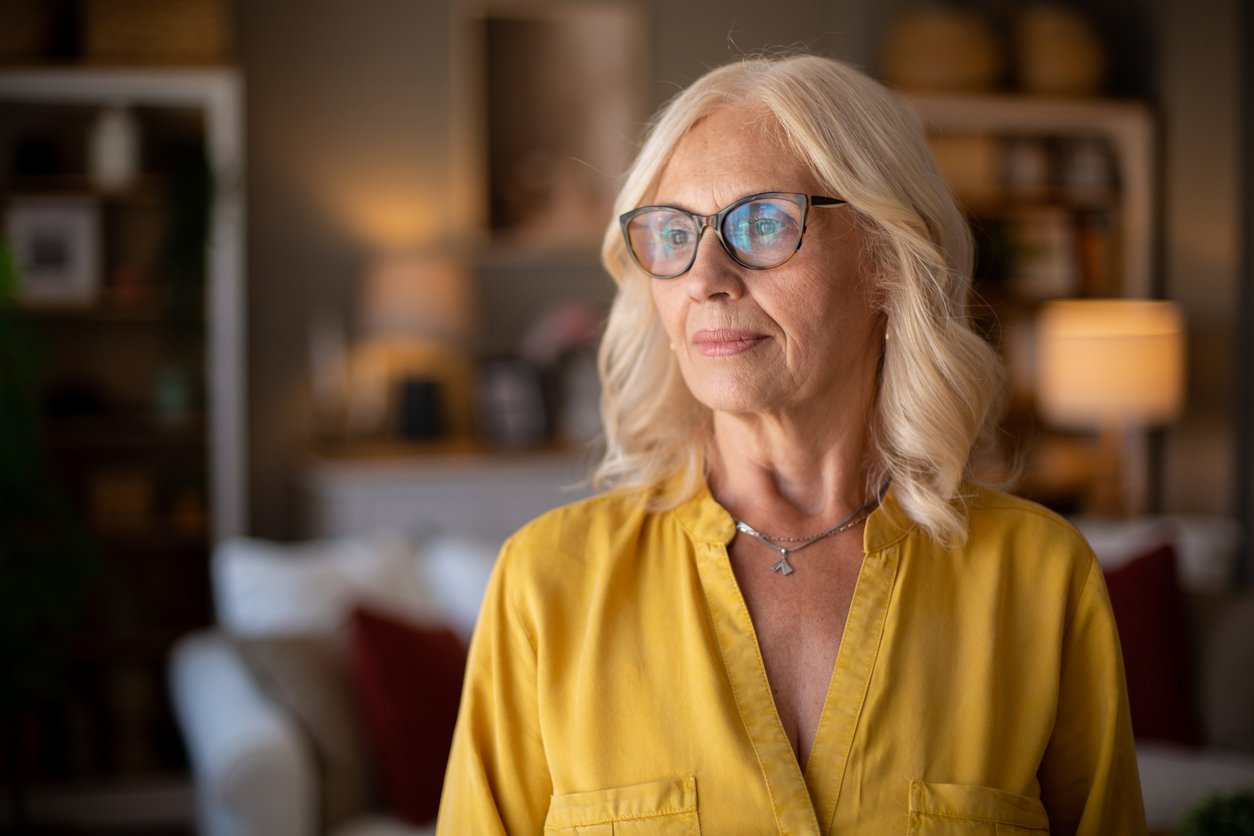 Elderly woman with white hair and glasses wearing a yellow blouse stands indoors, looking thoughtfully to the side. The background is softly lit with home decor and furniture visible.