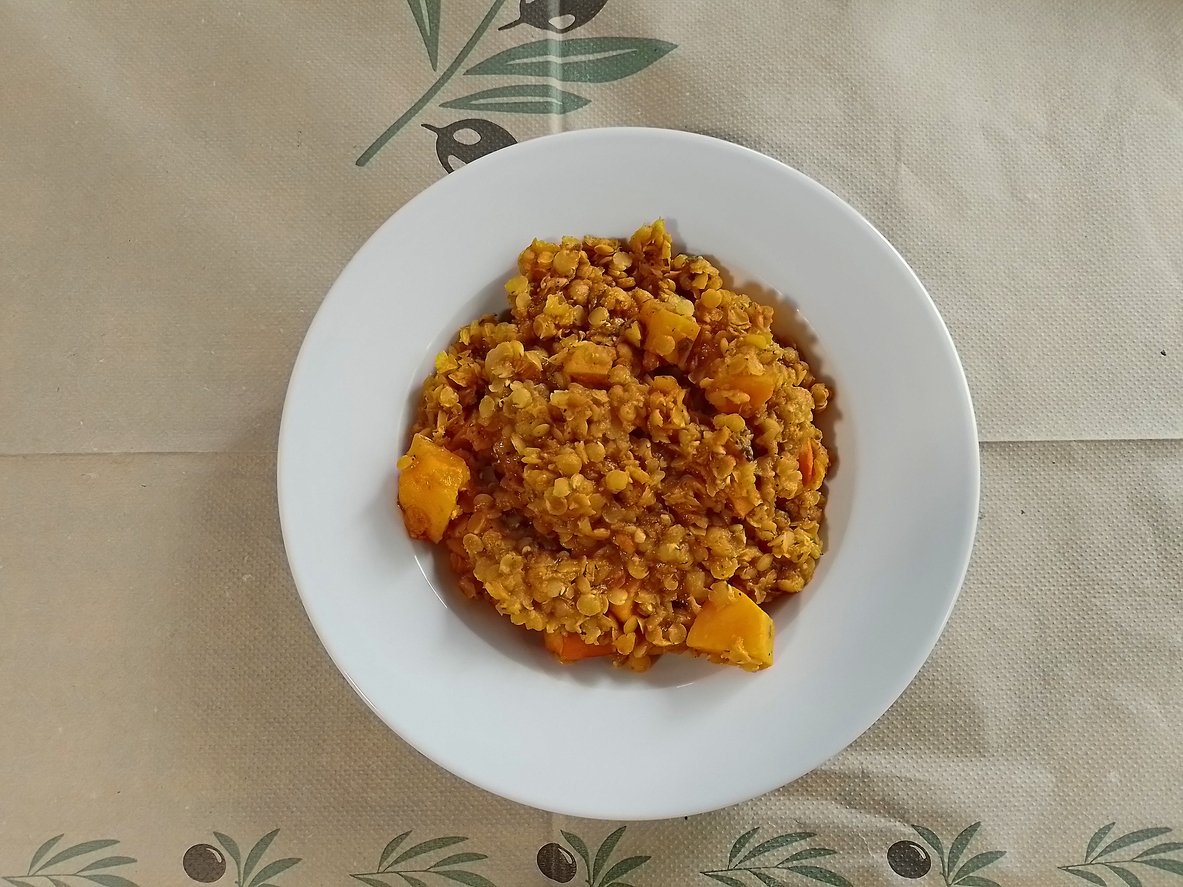 A white bowl filled with a lentil and vegetable stew, featuring visible chunks of orange squash, placed on a beige tablecloth with a green and black olive branch design.