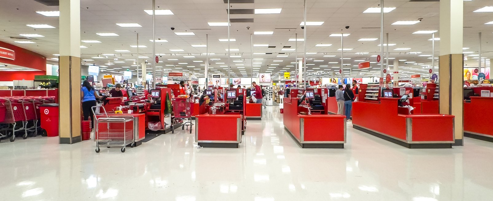 Wide view of a brightly lit retail store checkout area with red counters and several customers standing at registers; shopping carts are stacked on the left side of the image.