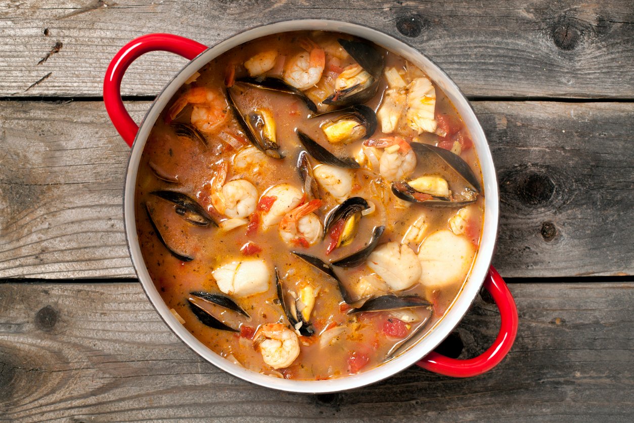 A pot of seafood stew with shrimp, mussels, scallops, and chunks of fish in a tomato-based broth, placed on a rustic wooden surface. The pot has red handles.