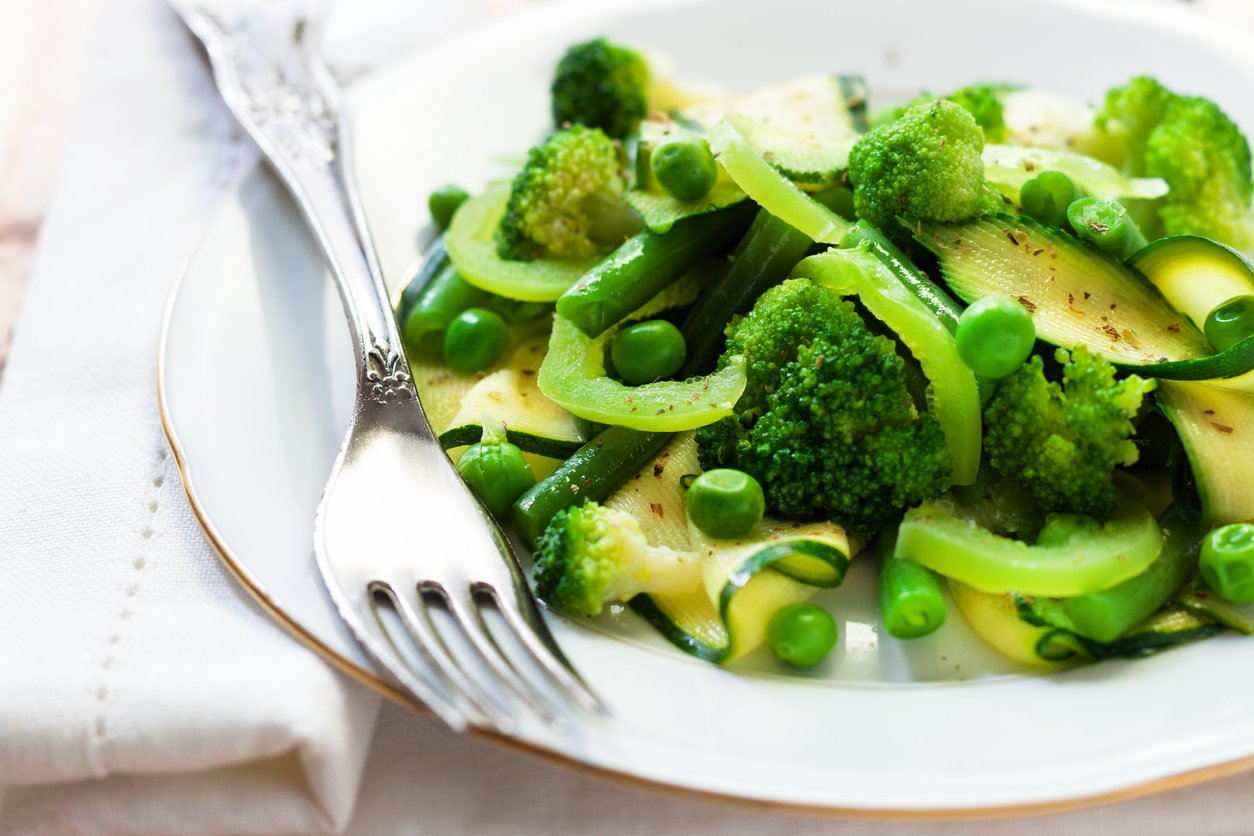 A white plate filled with a medley of green vegetables, including broccoli, peas, green beans, zucchini, and bell pepper slices, sits next to a silver fork on a white napkin.