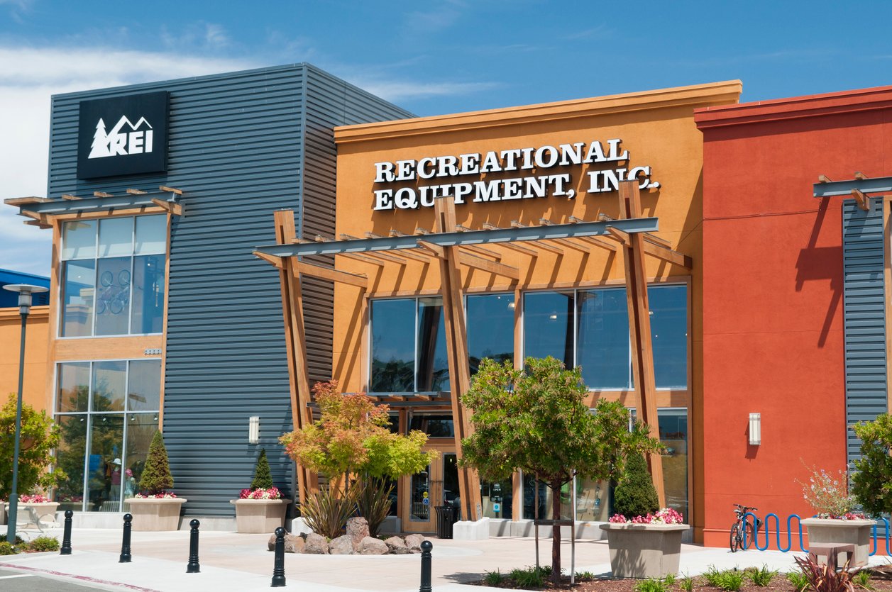 The exterior of an REI (Recreational Equipment, Inc.) store with orange and blue walls, large windows, wooden awning, and landscaped plants in front on a sunny day.