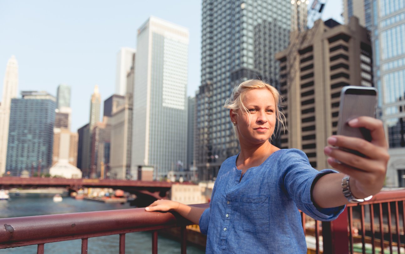 A woman in a blue shirt takes a selfie on a city bridge with tall skyscrapers and a river in the background on a sunny day.