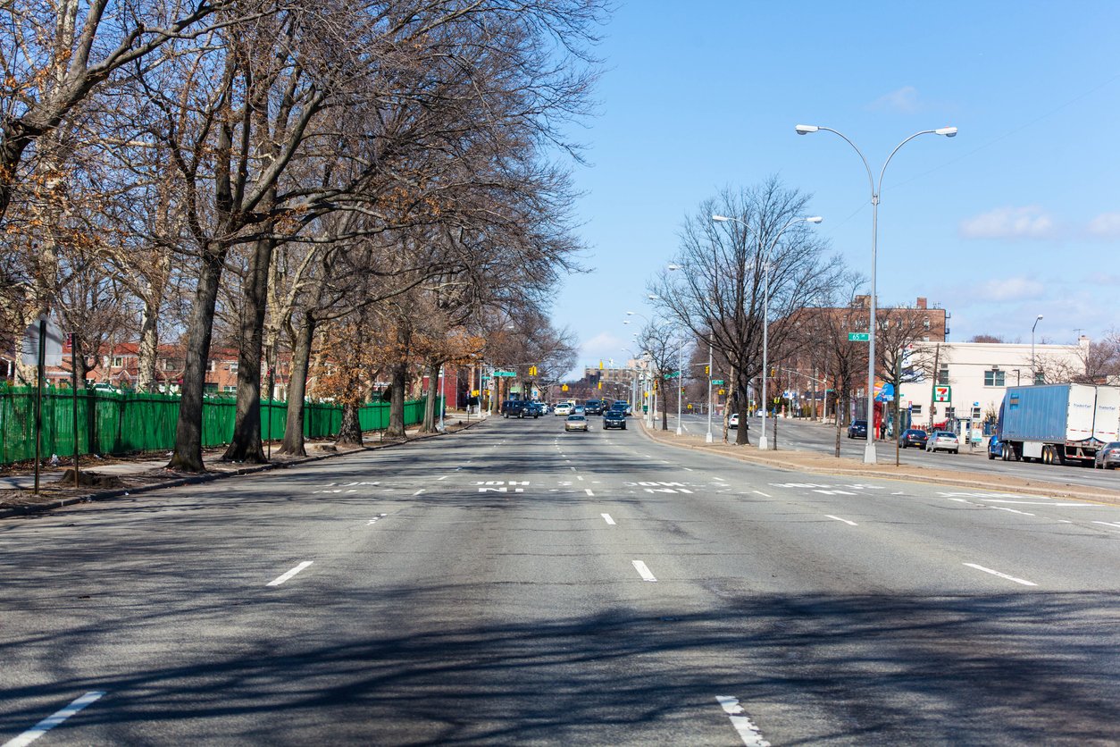 A wide, multi-lane urban street with a few cars driving, lined with leafless trees and green fencing on the left, under a clear blue sky. Buildings and traffic are visible in the distance.