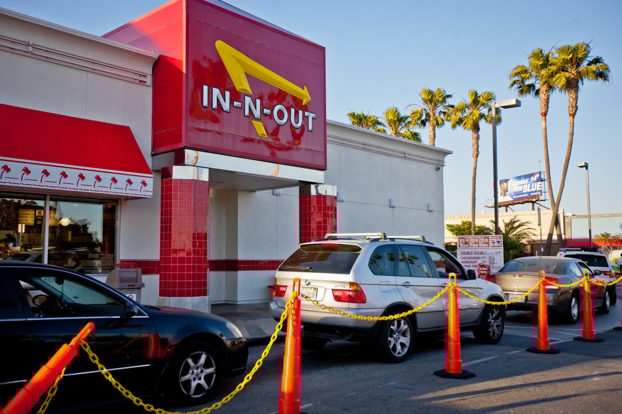 Cars lined up at the drive-thru of an In-N-Out Burger restaurant, with palm trees and a billboard in the background on a sunny day.