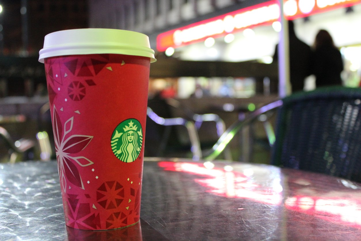 A red Starbucks holiday cup with a white lid sits on a reflective metal table outdoors, with blurred lights and chairs in the background, suggesting a café setting at night.
