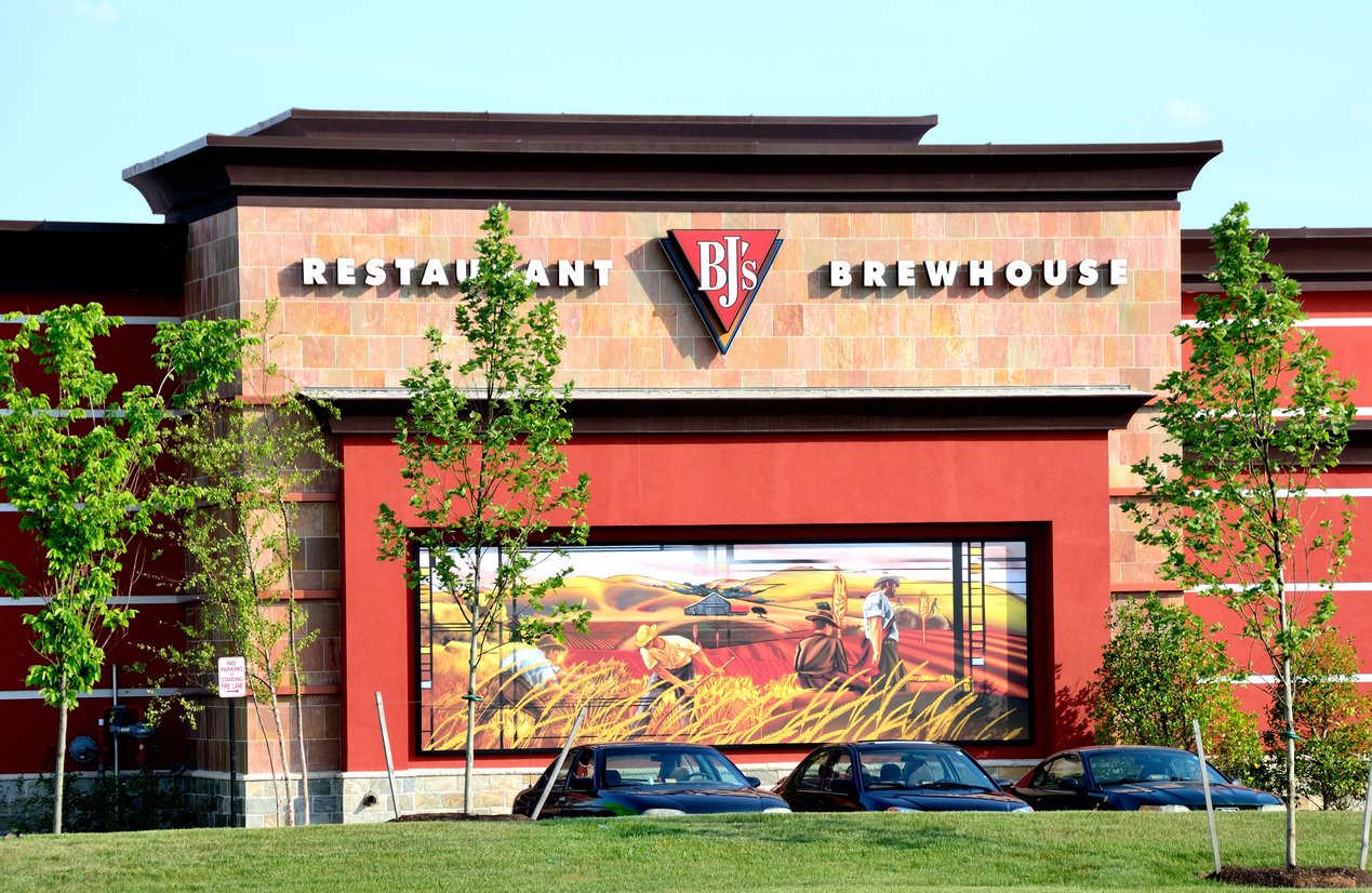 Exterior of BJ’s Restaurant Brewhouse with a mural and parked cars in front, trees partially obscuring the view, and a clear sky in the background.