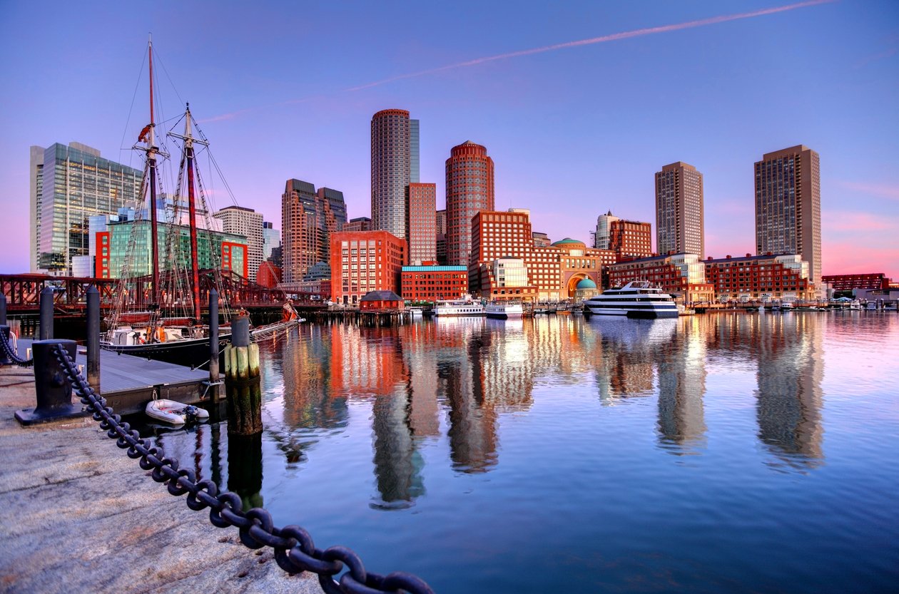 Boston harbor at sunset with modern and historic buildings, a docked sailboat, and yachts reflected in the calm water under a colorful sky.