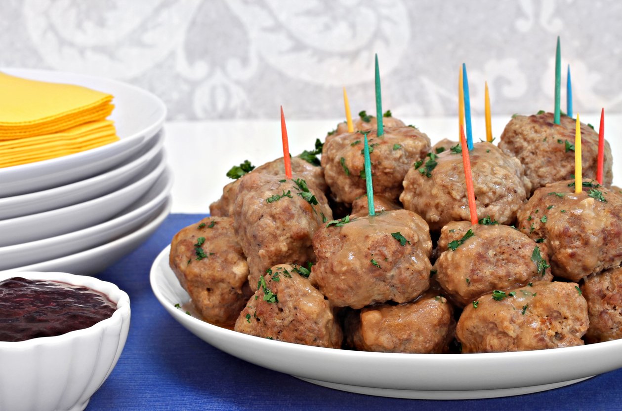 A white plate piled with Swedish meatballs, each held with a colorful toothpick, sits on a blue tablecloth. Nearby are stacked white plates, yellow napkins, and a small bowl of dark sauce.