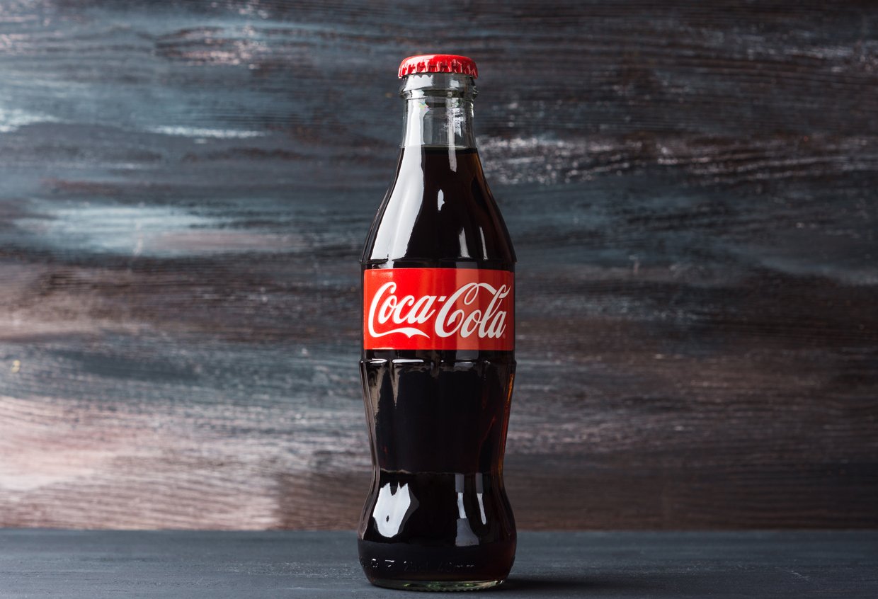 A classic glass bottle of Coca-Cola with a red cap and label stands on a dark surface against a rustic, textured background.