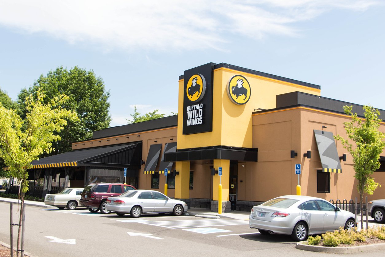 A Buffalo Wild Wings restaurant with yellow and black signage, several parked cars in front, and trees and blue sky in the background.