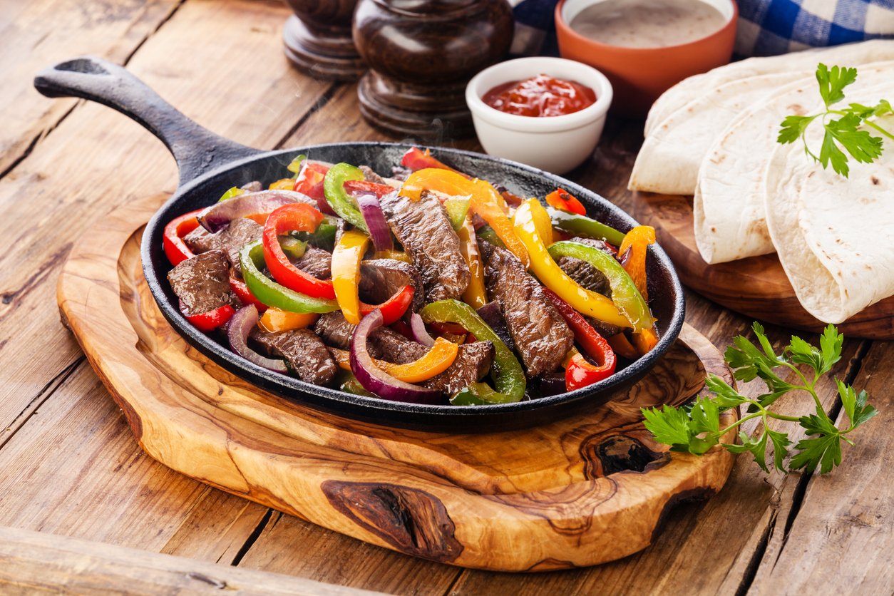 A sizzling skillet of beef fajitas with colorful bell peppers and onions on a wooden board, served with tortillas, salsa, sauce, and fresh parsley on the side.