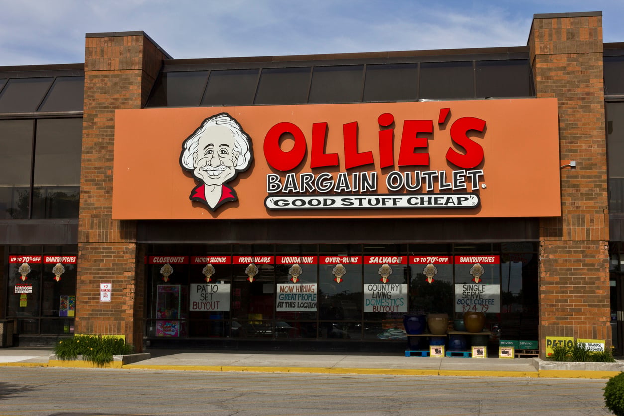 The storefront of Ollie’s Bargain Outlet features a large orange sign with a cartoon character and the slogan “Good Stuff Cheap.” The windows display various sale signs and product promotions.