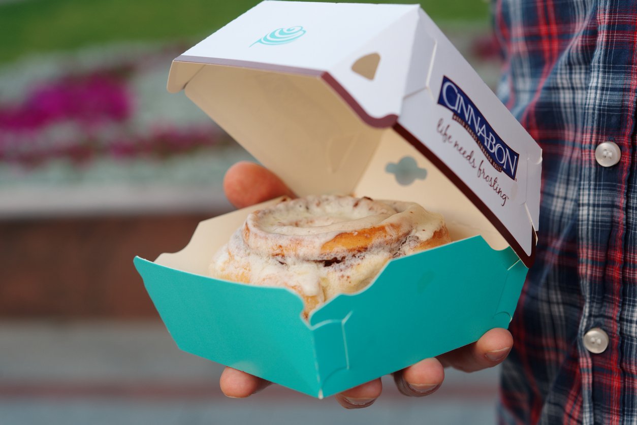 A person holding an open teal Cinnabon box with a frosted cinnamon roll inside. The person is wearing a plaid shirt and is standing outdoors, with blurred flowers in the background.
