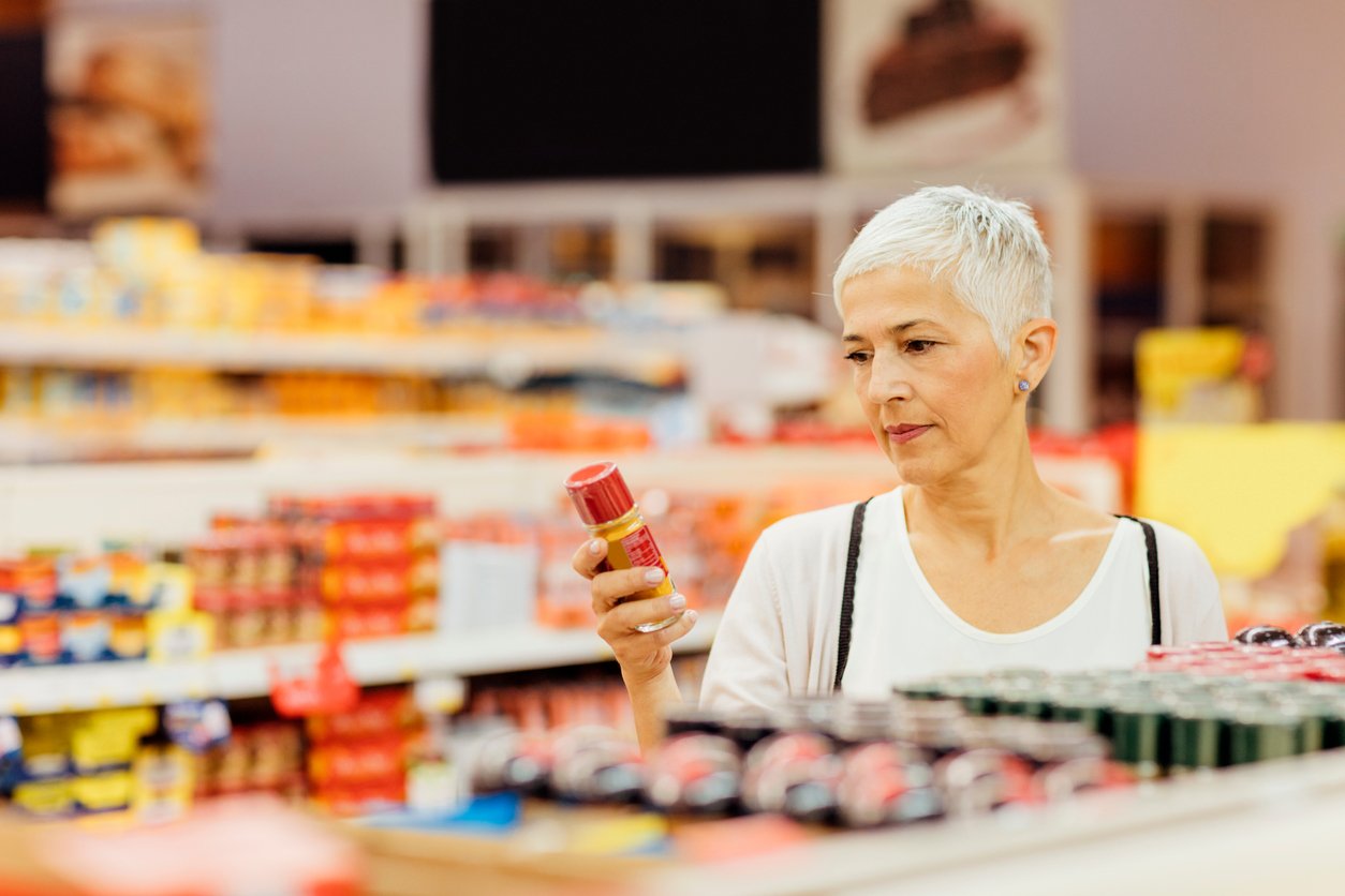 An older woman with short gray hair examines a spice jar while shopping in a grocery store aisle filled with various products.