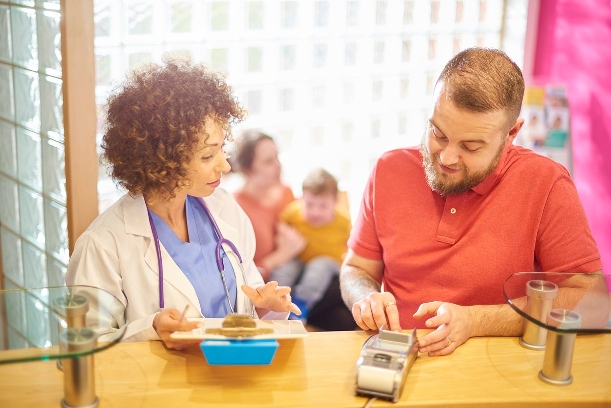 A doctor shows a clipboard to a man making a payment at a reception desk, while a woman and child wait in the background.