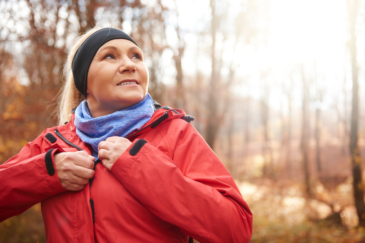 A woman outdoors in a forest during autumn zips up her red jacket, wearing a blue neck gaiter and black headband, with sunlight streaming through the trees in the background.