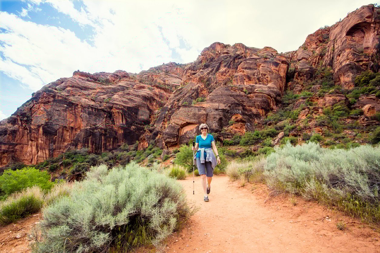 A person wearing sunglasses and hiking gear walks along a dirt trail surrounded by green shrubs, with tall, reddish rock formations rising in the background under a partly cloudy sky.