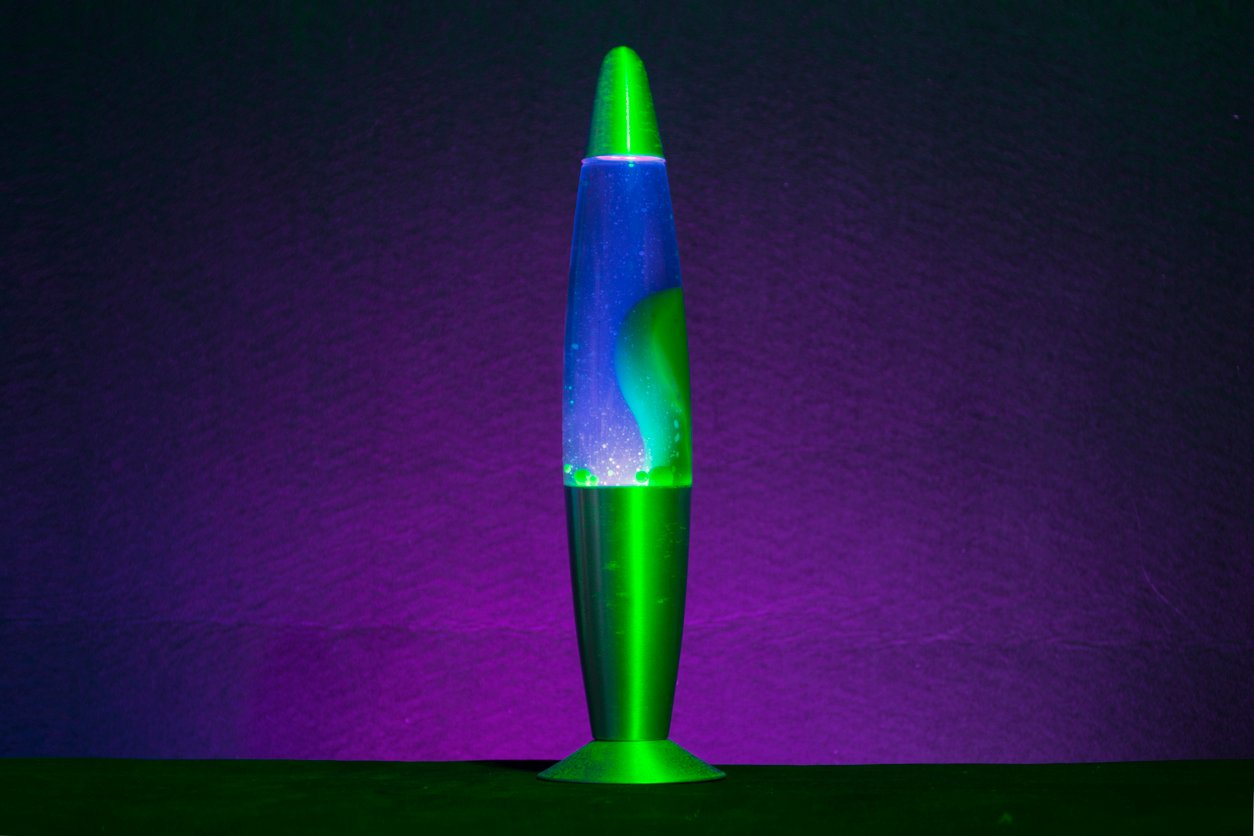 A green and blue lava lamp glows against a dark purple background, with swirling shapes visible inside the liquid.